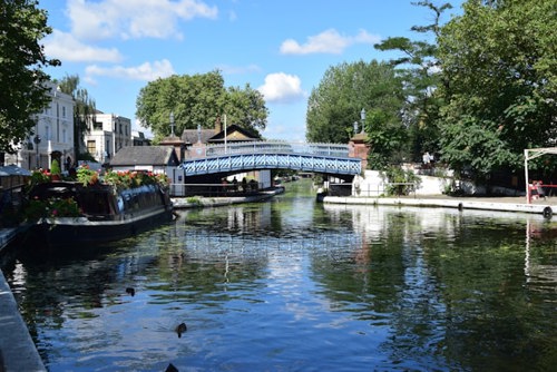 Uma fotografia panorâmica de barcos-casa em Little Venice, Londres, com céu azul e árvores verdes a rodear águas limpas.
