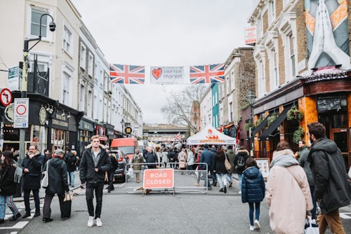 Fotografia em grande angular de pessoas a caminhar pelo Portobello Road Market. Bandeiras da Union Jack esvoaçam sobre um letreiro com o texto Your Local Market